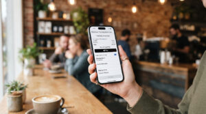 Person holding a modern smartphone displaying a secure e-commerce checkout page with Apple Pay, set against the softly blurred background of a stylish coffee shop.