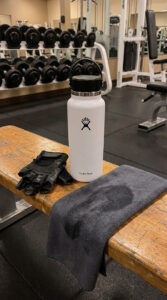 Hydro Flask 32 oz bottle placed on a gym bench beside workout gloves and a gym towel in a modern indoor gym.