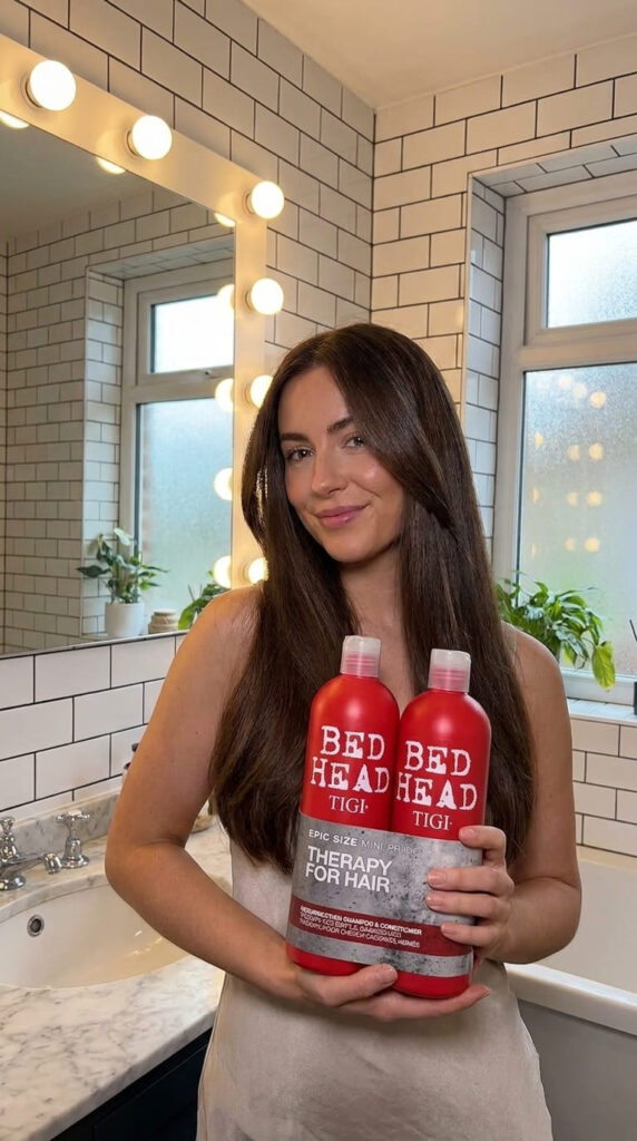 Person with healthy-looking shiny hair holding red Bed Head by TIGI Resurrection 750ml shampoo and conditioner bottles in a modern British bathroom.