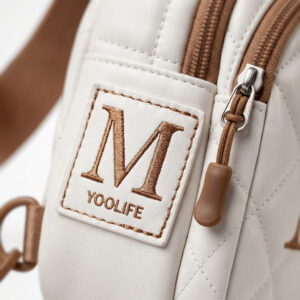 Close-up of the YOOLIFE sling bag showing its hand-stitched monogram initial patch and smooth nylon zipper pull.