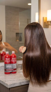 Person brushing through silky, freshly washed hair in front of a bathroom mirror, showing a smooth and glossy-looking finish.