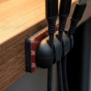Close-up of a multi-slot silicone cord clip with strong 3M adhesive backing attached to a desk and holding braided USB charging cables.